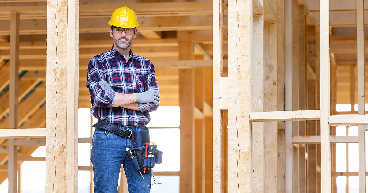 Home building standing in front of construction site with arms crossed and yellow hard hat